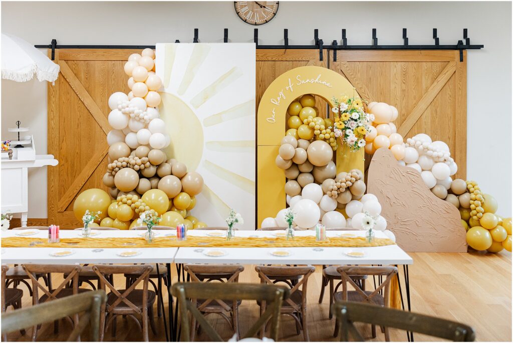 Sunny baby shower setup at Eagan Community Center with soft yellow tones, natural wood chairs, and floral arrangements in baskets.