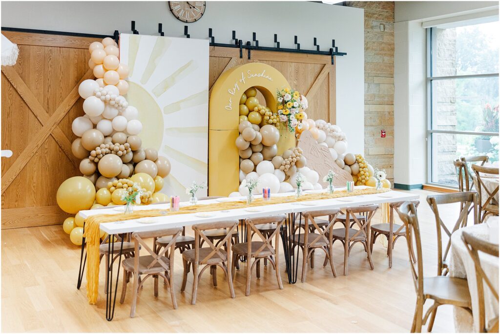 Sunny baby shower setup at Eagan Community Center with soft yellow tones, natural wood chairs, and floral arrangements in baskets.