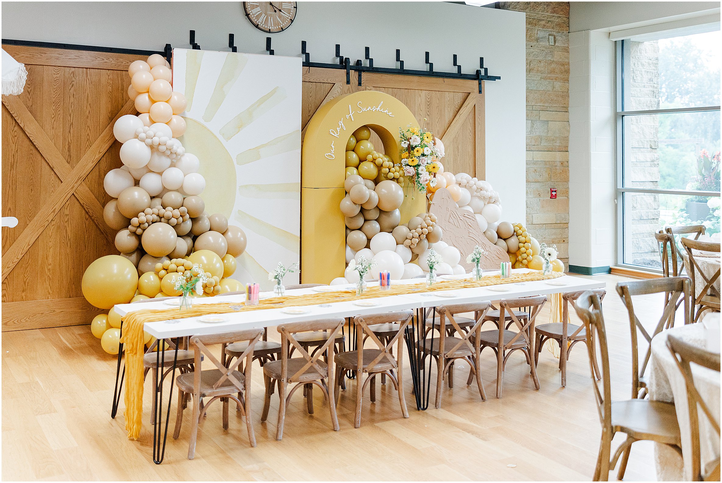 Sunny baby shower setup at Eagan Community Center with soft yellow tones, natural wood chairs, and floral arrangements in baskets.