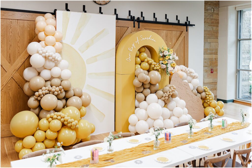 Sunny baby shower setup at Eagan Community Center with soft yellow tones, natural wood chairs, and floral arrangements in baskets.