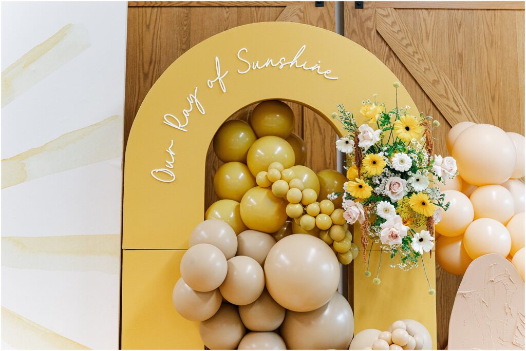 Sunny baby shower setup at Eagan Community Center with soft yellow tones, natural wood chairs, and floral arrangements in baskets.