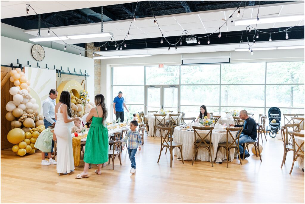 Sunny baby shower setup at Eagan Community Center with soft yellow tones, natural wood chairs, and floral arrangements in baskets.