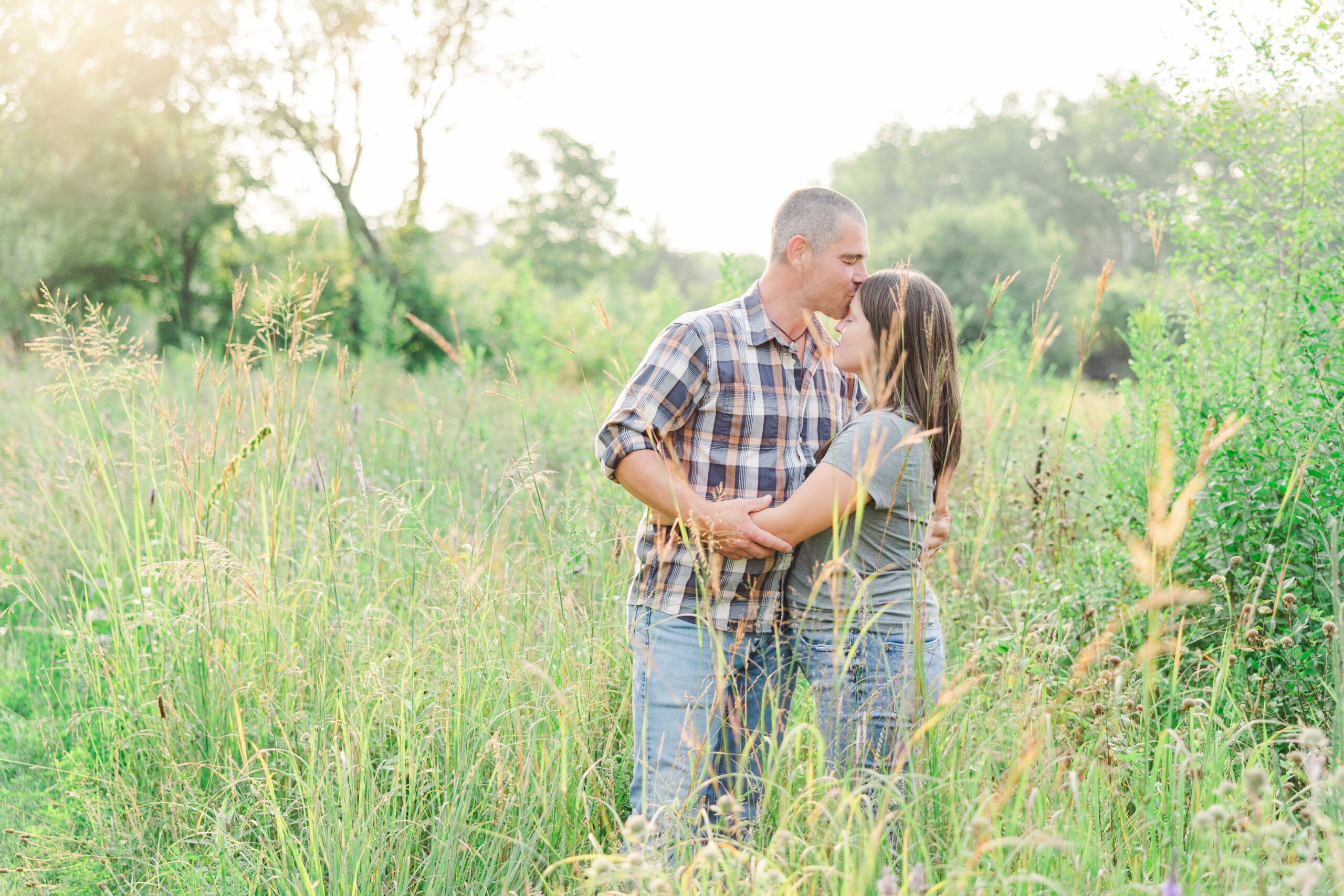 A couple standing in tall grass during a summer engagement session at Coon Rapids Dam Regional Park, with the golden hour sunlight filtering through the trees.